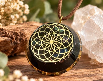 May include: A round black resin pendant featuring a gold-colored geometric flower of life pattern on the front surface. The pendant is suspended from a brown cord and contains embedded dark blue and purple stone fragments within the resin base.