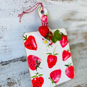 May include: A small, white, wooden cutting board with a strawberry pattern. The board has a red and white twine hanging loop. A real strawberry and sprigs of greenery are attached to the board.