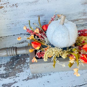 May include: A decorative fall centerpiece featuring a white, textured pumpkin atop a wooden trough. The trough is filled with colorful faux flowers and foliage in shades of orange, red, and yellow, creating a rustic autumn theme.