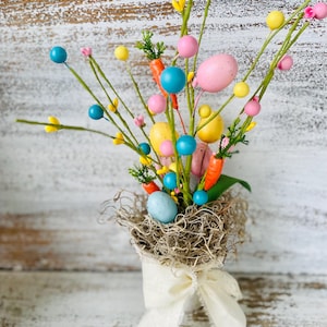 May include: A spring-themed floral arrangement in a white burlap-wrapped vase. The arrangement features pastel pink, yellow, and blue Easter eggs, carrots, and green sprigs.