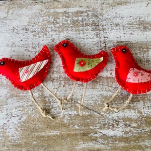 May include: Three handmade red felt bird ornaments with decorative fabric wings and button accents. Each bird has stitched details and twine legs, set against a rustic, weathered wooden background. These charming decorations add a touch of whimsy.