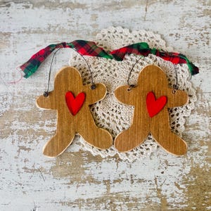 May include: Two wooden gingerbread man ornaments with red heart accents. The ornaments are brown with a distressed finish and are suspended by black wire. A red, green, and black plaid ribbon is attached for hanging. The ornaments are placed on a cream-colored doily.