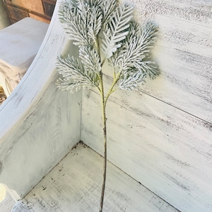 May include: A frosted green twig spray with a brown stem. The twig spray is on a white painted wooden surface.