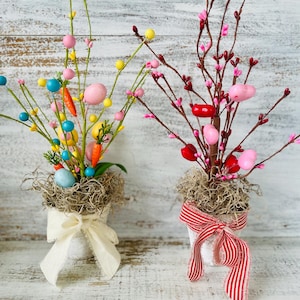 May include: Two small white pots with a nest of dried moss and colorful Easter eggs. One pot has a white ribbon and the other has a red and white striped ribbon.