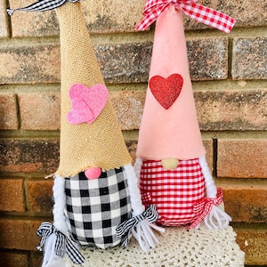 May include: Two Valentine's Day gnomes, one with a burlap hat and black and white checkered fabric, and the other with a pink hat and red and white checkered fabric. Both gnomes have white yarn hair and a pink nose.