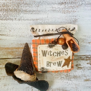 May include: A small, orange and white plaid fabric bag with the words "Witches Brew" and a star. The bag is tied with twine and has a wooden scoop inside. A black witch's hat is next to the bag.