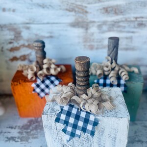 May include: Three decorative wooden pumpkins in orange, white, and green. Each pumpkin has a wooden stem, burlap ribbon, and a black and white checkered leaf accent. Rustic fall decor.