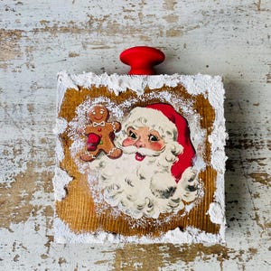 May include: A Christmas-themed bottle stopper featuring a vintage Santa Claus illustration with a gingerbread man. The stopper has a red knob and is set on a wooden block with a textured, snowy border.