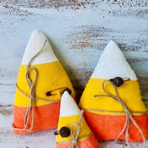 May include: Three handmade candy corn decorations. Each has a white, yellow, and orange color scheme. They are tied with twine and have a small bell and safety pin attached. The decorations are placed on a distressed white wooden surface.