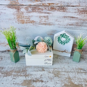 May include: A decorative arrangement featuring a stack of white books with the text "Our Happy Place", a small house-shaped sign with a wreath design, and two small green vases with faux greenery. The arrangement includes faux flowers in shades of peach and green.