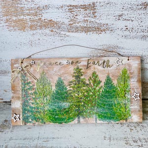 May include: Wooden sign with the words "Peace on Earth" and a row of green Christmas trees. The sign has a rustic, distressed look with star-shaped embellishments and a twine hanger. The background is a light brown color.