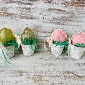 May include: Four small white pots with green grass and fabric Easter eggs. The eggs are green with white polka dots, green and white gingham, pink, and pink and white stripes.