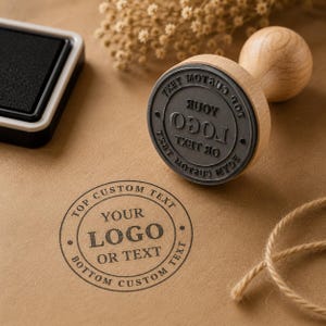 May include: A round wooden rubber stamp with a handle, featuring a black rubber base with the text YOUR LOGO OR TEXT. A matching black ink pad is nearby, and a circular impression of the stamp is visible on the brown paper surface below.