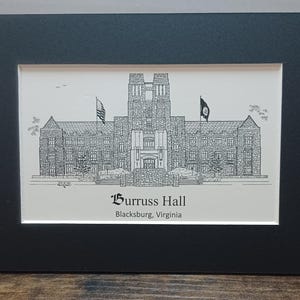 May include: Black framed print of Burruss Hall in Blacksburg, Virginia. The detailed black and white illustration depicts the building's facade, flags, and surrounding trees. The text "Burruss Hall" and "Blacksburg, Virginia" are below the image.