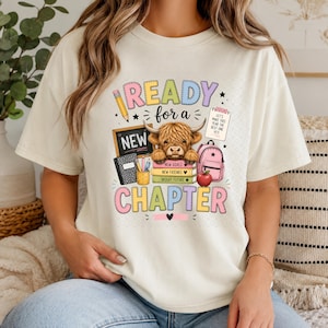 Może przedstawiać: Kremowy t-shirt z grafiką przedstawiającą szkocką wyżynną krowę na stosie książek z napisami New Goals, New Friends i Bright Future. Tekst brzmi Ready for a New Chapter, a obok znajduje się ołówek, tablica szkolna oraz plecak.