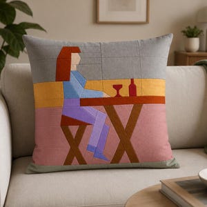 May include: A square throw pillow featuring a geometric fabric appliqué design of a person sitting at a table with a glass and bottle. The scene uses blocks of gray, yellow, pink, blue, orange, and brown fabric with visible stitching lines.