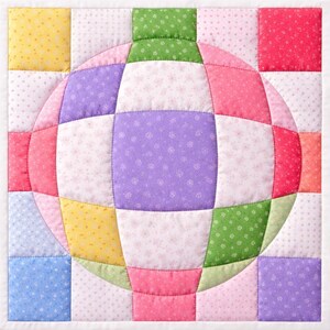 May include: A square quilted wall hanging featuring a central sphere design. The sphere is composed of various fabric squares in shades of pink, purple, green, and yellow, with a white background. The quilt is machine-stitched with a white border.