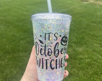May include: A clear plastic tumbler with a glittery iridescent finish and a black vinyl decal that reads "It's October Witches". The tumbler has a black straw.