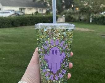 May include: A clear plastic tumbler filled with green glitter and a purple ghost design. The tumbler has a clear lid and a gray straw. The text "Boo Season" is around the ghost. The tumbler is held in front of a green lawn.