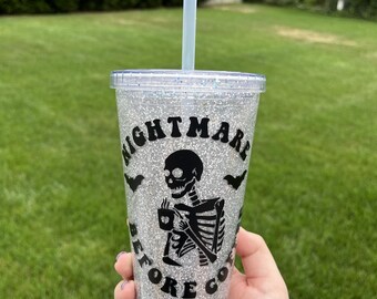 May include: Glittery silver tumbler with a black skeleton graphic holding a coffee cup. The text "NIGHTMARE BEFORE COFFEE" is printed around the skeleton. Includes a clear straw. Perfect for Halloween or spooky season.