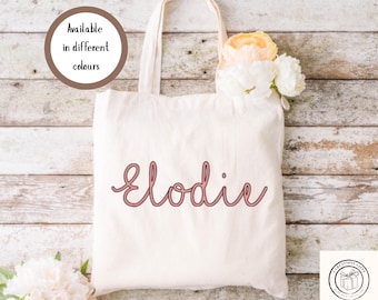 May include: Cream-colored tote bag with the name "Elodie" in pink script. The bag has a simple design with long handles and is decorated with flowers. Text in a circle reads "Available in different colours".