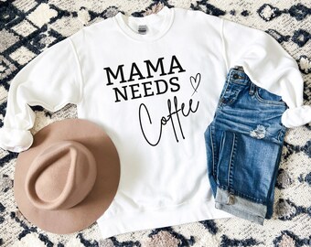 May include: White sweatshirt with black text that reads "Mama Needs Coffee" with a small heart symbol. The sweatshirt is laying on a white and gray patterned rug with a brown hat and blue jeans.
