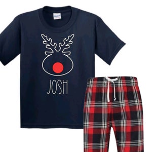 May include: Navy blue t-shirt with a white reindeer design and the name "Josh" printed on the front. The shirt is paired with red and black plaid pajama pants.