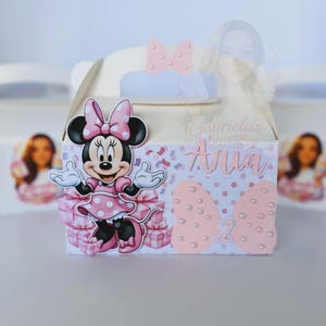 May include: A white party favor box featuring a Minnie Mouse design. The box has a pink bow and the name "Anna" printed on it. The number "2" is also displayed. The box is decorated with pink polka dots and confetti.