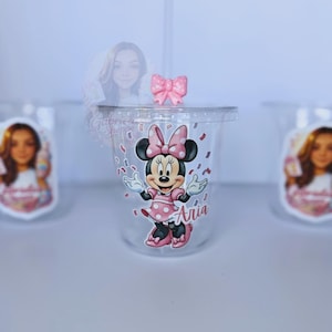 May include: A clear plastic cup with a lid and straw, featuring a Minnie Mouse design. The cup has a pink bow on top and a Minnie Mouse sticker with the name "Aria". Confetti-like decorations are scattered around the cup.