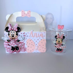 Könnte beinhalten: Eine weiße Partygeschenkbox, ein durchsichtiger Plastikbecher und ein Deckel, alle im Minnie-Mouse-Design. Die Box und der Becher zeigen Minnie-Mouse-Bilder, Schleifen und den Namen "Aria". Die Box hat eine rosa Schleife oben.