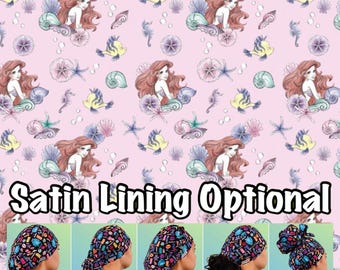 May include: A pink fabric with a repeating pattern of Ariel, the Little Mermaid, with seashells, starfish, and seahorses. The fabric is for making surgical caps. The text "Satin Lining Optional" is at the bottom of the image. Five different styles of surgical caps are shown below the text: Euro, Ponytail, Bouffant, Tie Back, and High Bun.