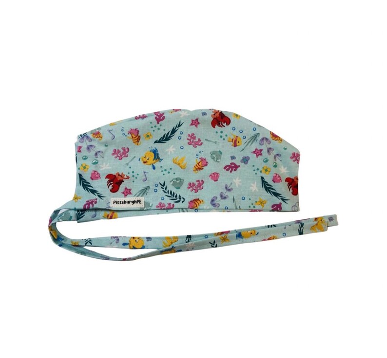 May include: A light blue scrub cap featuring a repeating pattern of colourful cartoon fish, crabs, seashells, and seaweed. The fabric head covering includes long ties for adjustment and a small white rectangular tag labelled PittsburghPE.