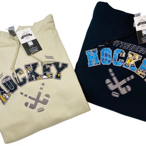 May include: Two hooded sweatshirts with a Pittsburgh Hockey design. One is beige with black and white lettering and the other is navy blue with blue and white lettering.