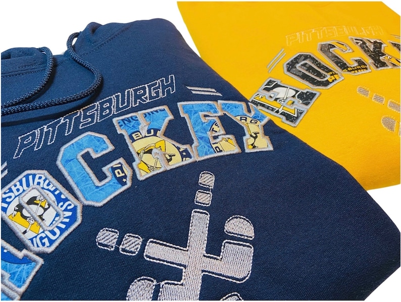 May include: A navy blue hoodie with a white embroidered design that says "Pittsburgh Hockey" with a penguin logo. The hoodie has a drawstring hood and a kangaroo pocket.