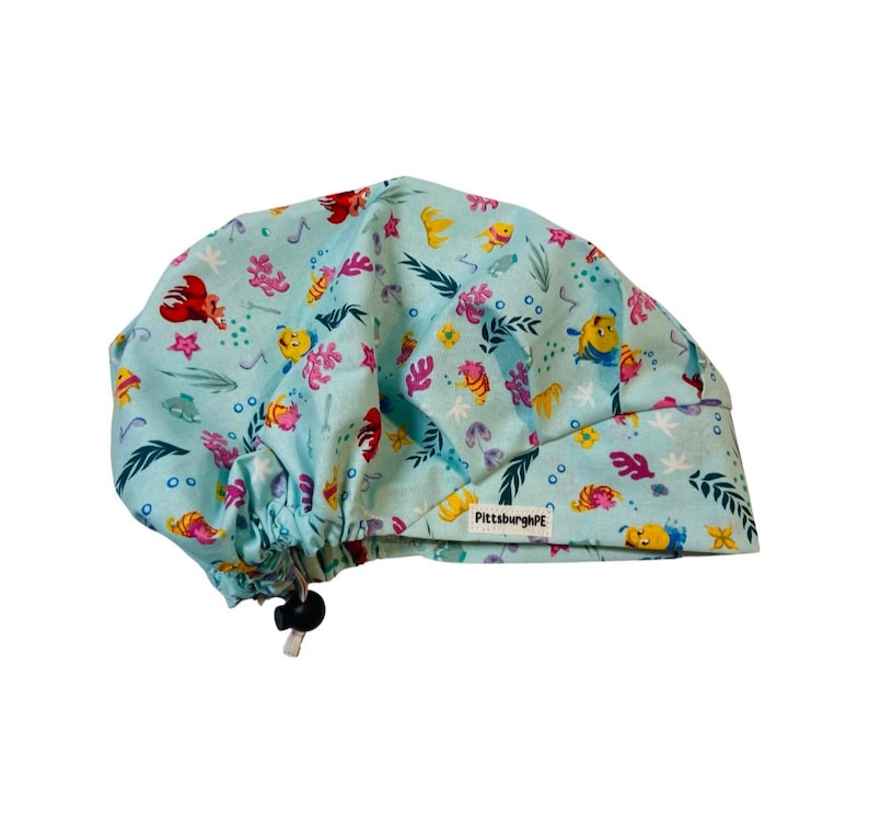 May include: A light blue scrub cap featuring a repeating pattern of colourful cartoon fish, crabs, and coral reef elements. The cap includes a black adjustable toggle cord at the back and a small white fabric tag labelled PittsburghPE.