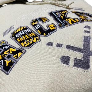 May include: A cream-colored hoodie with a large embroidered graphic of the word "ROCKS" in black and yellow. The graphic is made up of smaller text and images. The hoodie also has a smaller embroidered graphic of a hockey stick on the right side.