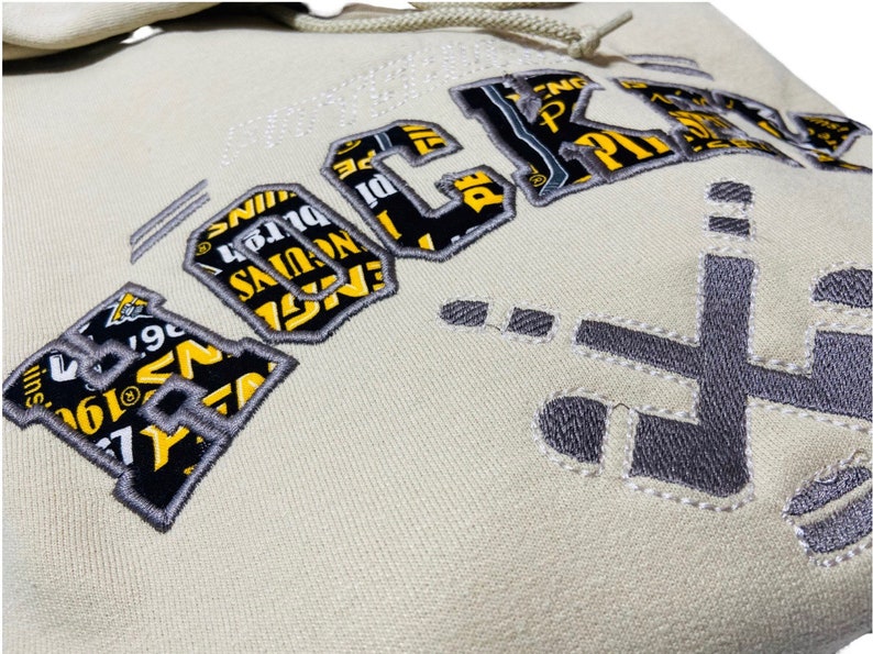 May include: A cream-colored hoodie with a large embroidered graphic of the word "ROCKS" in black and yellow. The graphic is made up of smaller text and images. The hoodie also has a smaller embroidered graphic of a hockey stick on the right side.