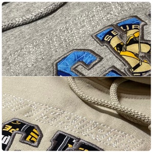 May include: Close-up of two gray hooded sweatshirts with embroidered logos. The top sweatshirt has a blue and yellow logo with the text "SEATTLE" and the bottom sweatshirt has a black and yellow logo with the text "SEATTLE".