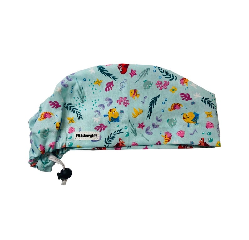 May include: A light blue scrub cap featuring a repeating pattern of yellow fish, pink coral, green seaweed, and bubbles. The fabric cap includes a white rectangular tag with the black text PittsburghPE and a black adjustable cord lock closure.