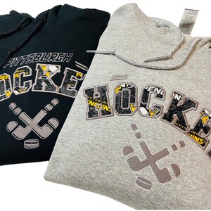May include: Two hooded sweatshirts, one black and one gray, with a Pittsburgh Penguins hockey themed design. The design features the word "HOCKEY" with a hockey stick and puck graphic.