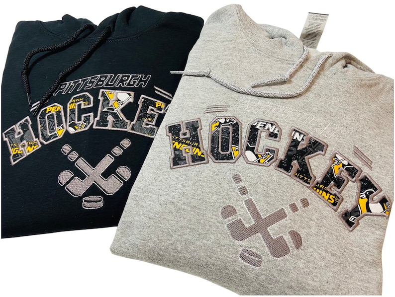 May include: Two hooded sweatshirts, one black and one gray, with a Pittsburgh Penguins hockey themed design. The design features the word "HOCKEY" with a hockey stick and puck graphic.
