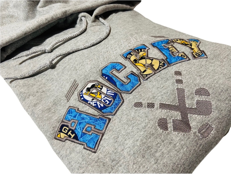 May include: Gray hooded sweatshirt with a blue and yellow embroidered design that says "HOCKEY" with a hockey stick and puck graphic. The design is made up of different hockey team logos.