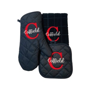 May include: A set of three black kitchen accessories with quilted fabric. The set includes a potholder, an oven mitt, and a kitchen towel. All three items feature the name "Cofield" embroidered in white with a red "C" initial.