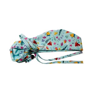 May include: A light blue scrub cap featuring a colourful pattern of cartoon fish, crabs, and underwater plants. The cap has a gathered back, long fabric ties for adjustment, and a small white tag with the text PittsburghPE on the side.