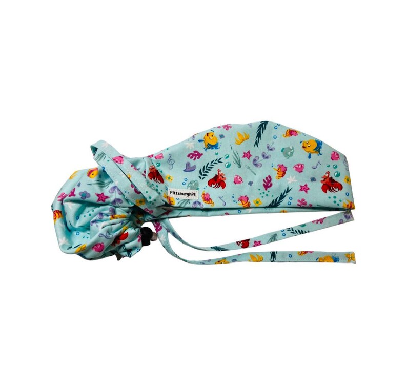 May include: A light blue scrub cap featuring a colourful pattern of cartoon fish, crabs, and underwater plants. The cap has a gathered back, long fabric ties for adjustment, and a small white tag with the text PittsburghPE on the side.