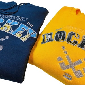 May include: Two sweatshirts, one navy blue and one yellow, with the text "Pittsburgh Hockey" embroidered on the front. The text is made up of smaller patches of fabric with different designs.