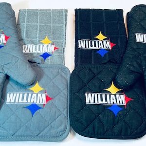 May include: Set of two oven mitts and two pot holders in gray and black with a quilted design. Each item features a yellow, blue, and red star with the name "William" embroidered on it.