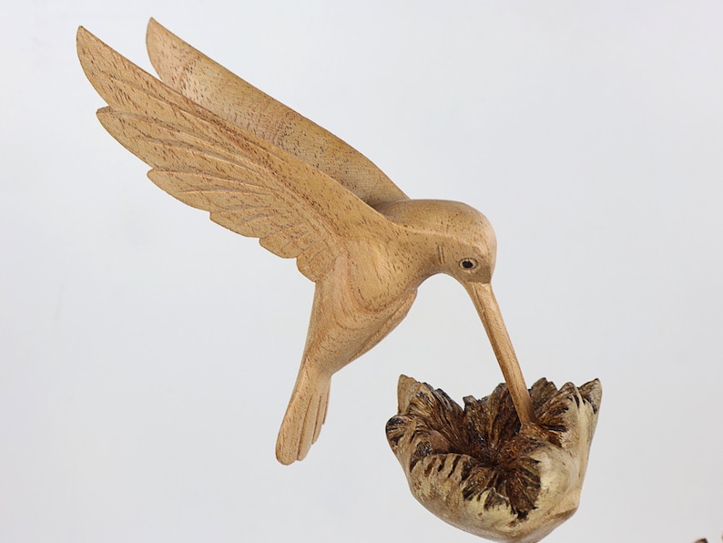 May include: A wooden hummingbird sculpture with its beak in a flower. The sculpture is hand-carved and painted in natural wood tones.