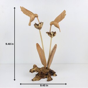 May include: A wooden sculpture of two hummingbirds perched on a flower with leaves. The sculpture is made of natural wood and has a rustic finish. It measures 9.48 inches tall and 9.45 inches wide.