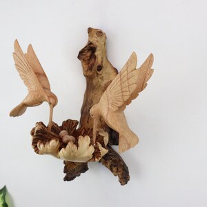 May include: A wooden sculpture of two hummingbirds perched on a branch with a nest of eggs. The birds are carved in detail and have a natural wood finish.